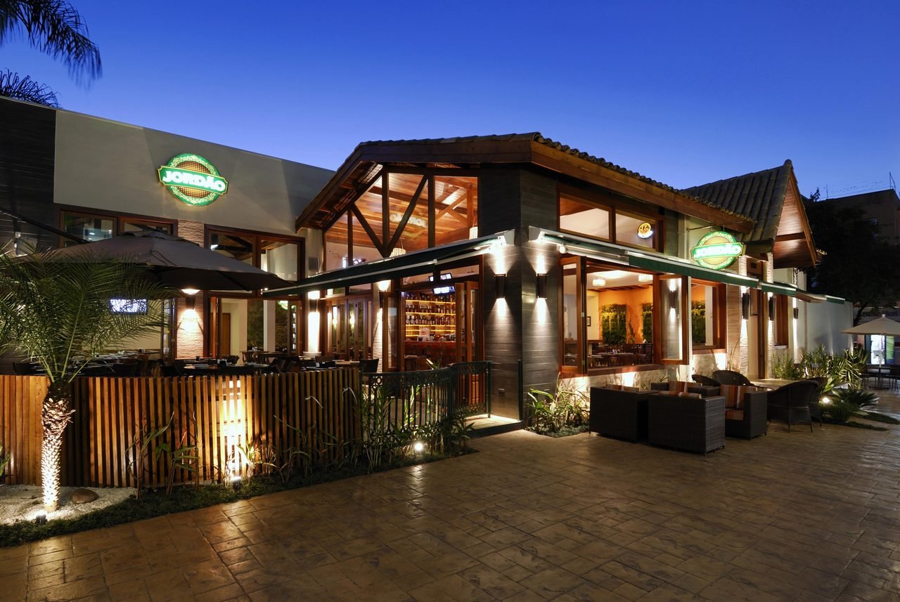 Fachada iluminada de um restaurante ao entardecer, com terraco, mesas e palmeiras, na zona leste de Sao Paulo